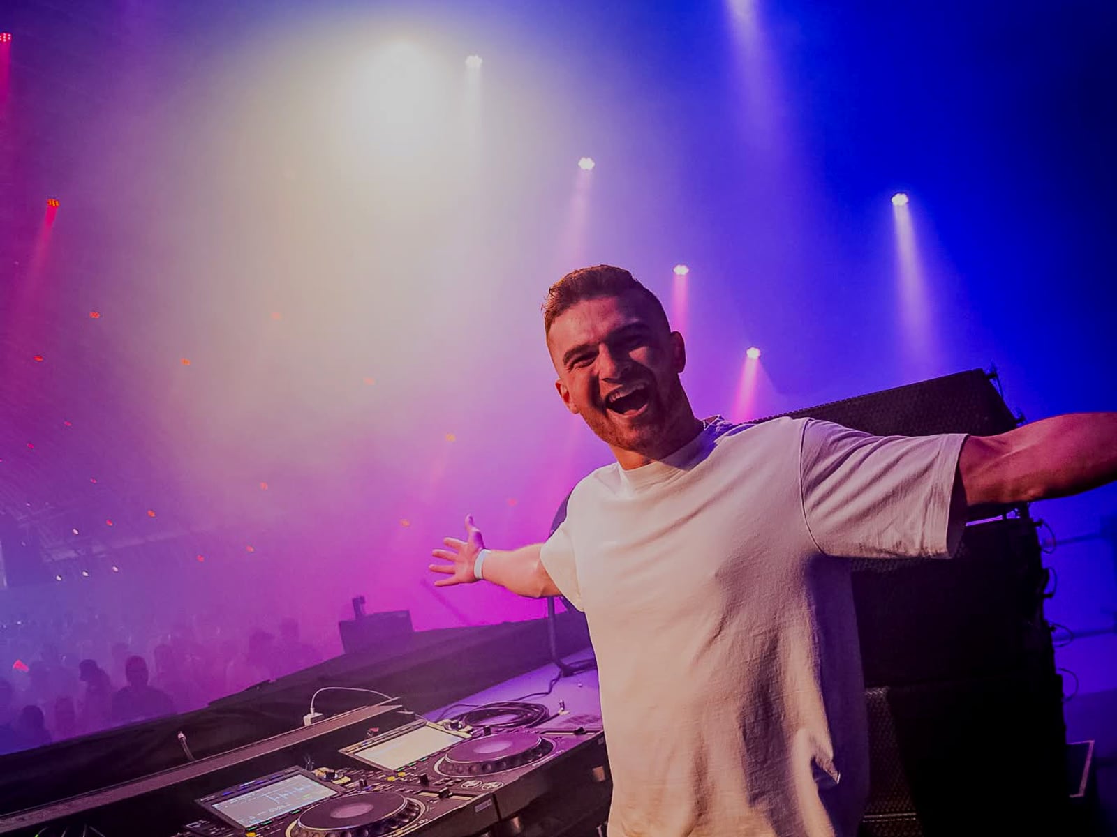 A-RIZE with both arms spread wide behind the decks under purple and pink light