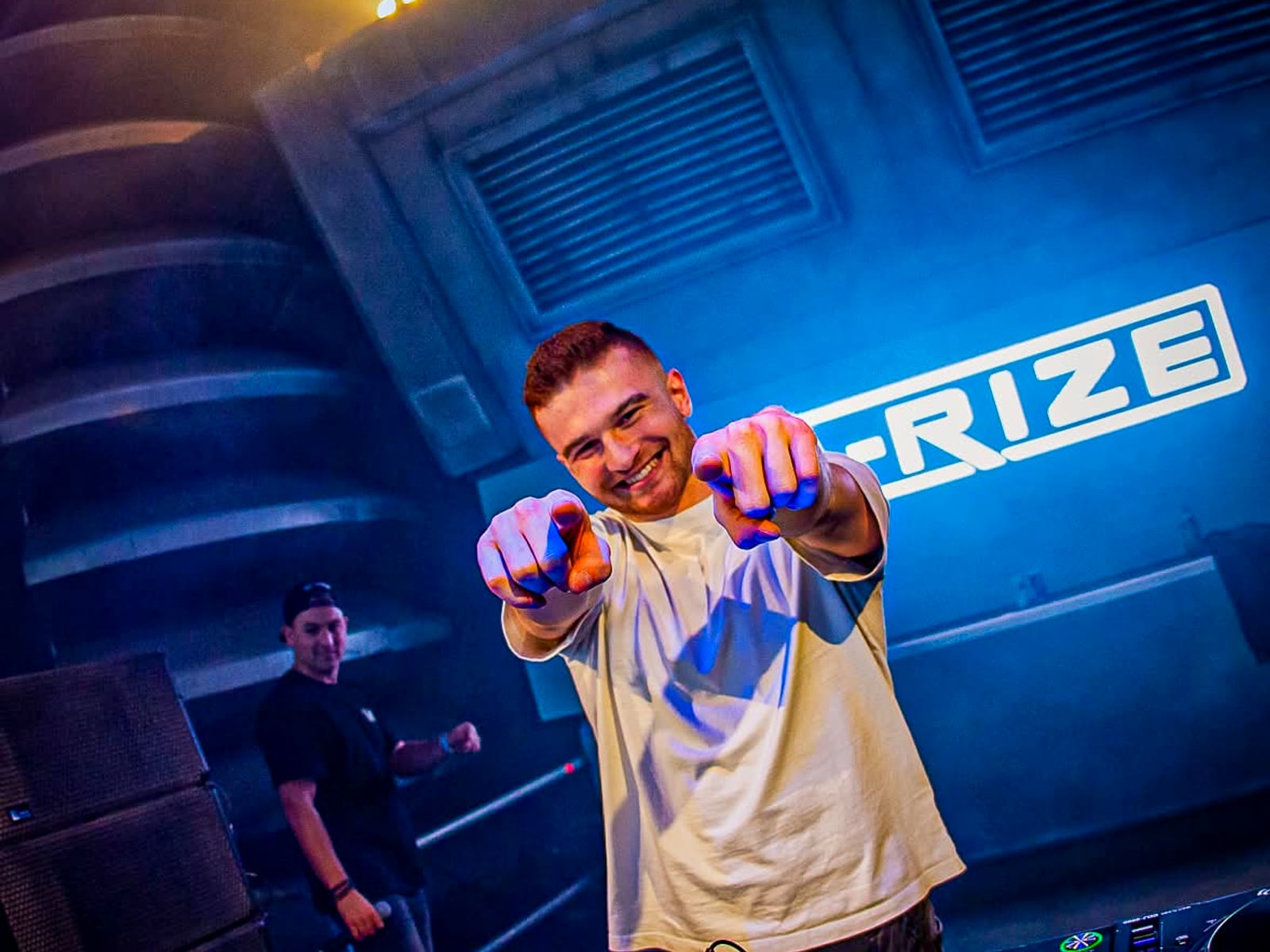 A-RIZE pointing at the camera from behind the CDJs, the A-RIZE logo lit on the wall behind him